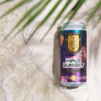 A Maui Sunset POG seltzer can on wet sand as sea foam rolls in, framed by a palm frond and hibiscus flowers
